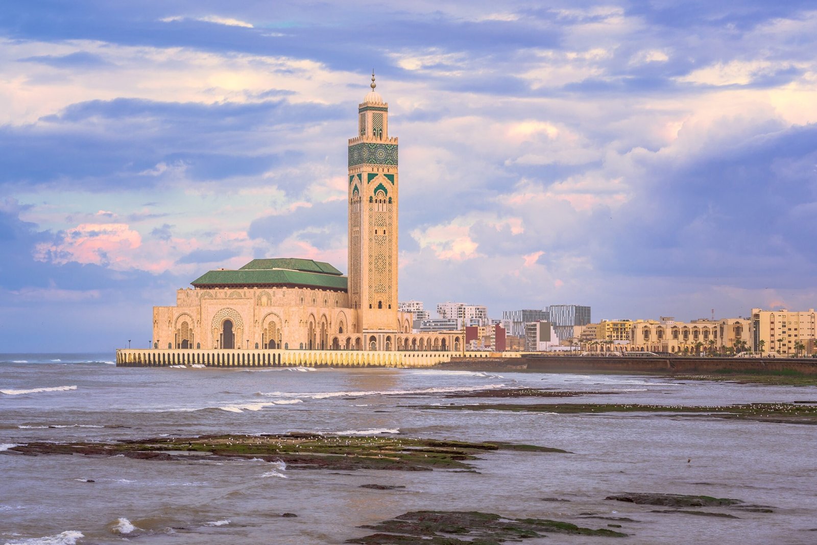 A wide landscape view of the majestic Hassan II Mosque standing on the Atlantic Ocean shoreline in Casablanca under a dramatic cloudy sky, explored during a private tour of Morocco.