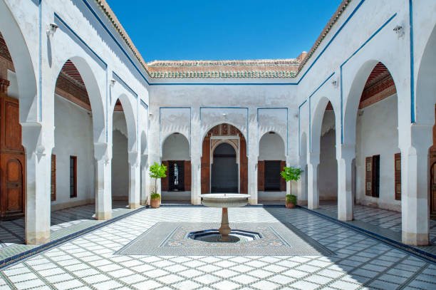 Symmetrical view of a traditional Moroccan riad courtyard with a central marble fountain in Bahia Palace in Marrakech, intricate zellij tile flooring, and white arched colonnades under a blue sky, experienced on a private tour of Morocco.