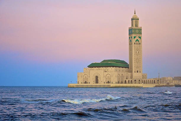 The monumental Hassan II Mosque built directly over the Atlantic Ocean in Casablanca, featuring its iconic tall minaret and green-tiled roofs under a pastel sunset sky during a private tour of Morocco.