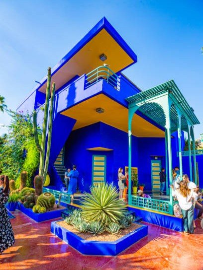 Majorelle Garden Blue Villa Marrakech | Private Tours of Morocco - Rafia Morocco Tours - Rafia Morocco Tours Bright cobalt blue and vivid yellow Cubist villa surrounded by exotic cacti and succulents at the Majorelle Garden, filled with visitors during a private tour of Morocco.