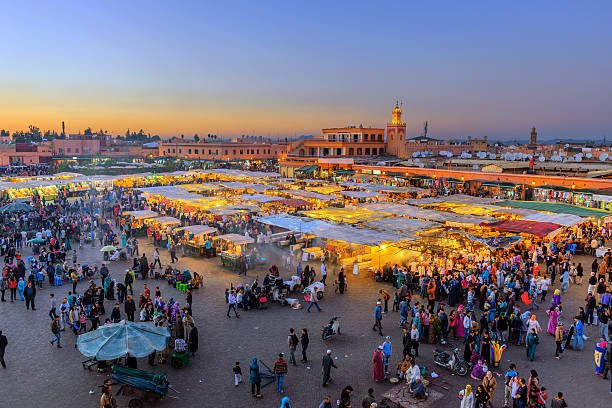An elevated high-angle view of the bustling Jemaa el-Fnaa square in Marrakech at sunset, filled with crowds and illuminated food stalls under a twilight sky during a private tour of Morocco.