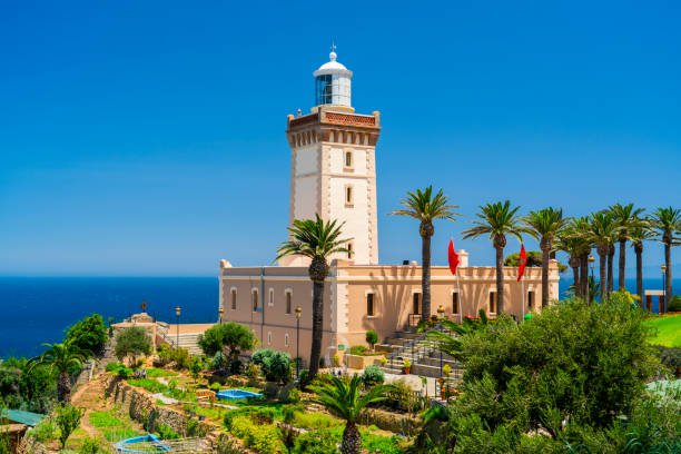 The historic Cape Spartel Lighthouse standing on a lush green cliffside lined with palm trees overlooking the deep blue Atlantic Ocean near Tangier, visited on a private tour of Morocco.