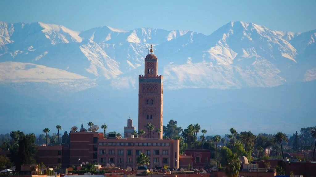 A wide cityscape view of Marrakech featuring the sandstone minaret of the Koutoubia Mosque rising above city buildings and palm trees, framed by the massive, snow-capped peaks of the High Atlas Mountains under a clear blue sky.