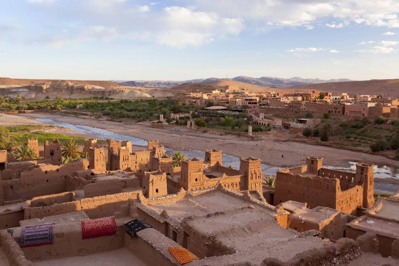 Ait Benhaddou Kasbah and River Valley | Private Tours of Morocco - Rafia Morocco Tours - Rafia Morocco Tours High-angle panoramic view of the ancient earthen clay kasbahs of Ait Benhaddou overlooking a wide, winding river valley and palm trees, with a modern village in the background, captured during a customized private tour of Morocco.