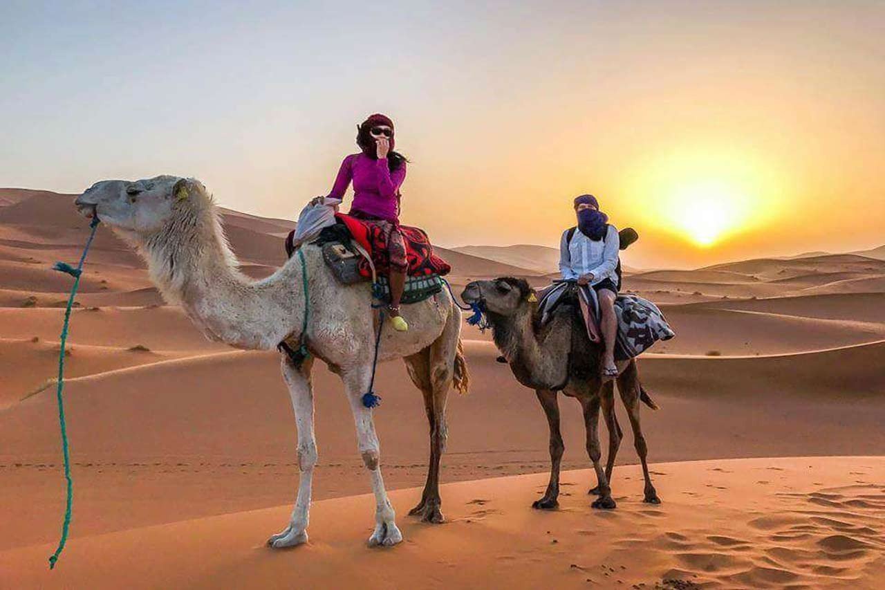 Sunset Sahara Camel Trek | Private Tours of Morocco - Rafia Morocco Tours - Rafia Morocco Tours Two travelers with protective headscarves riding camels across the vast orange sand dunes of the Sahara Desert at sunset, experiencing a tailored camel trek during a private tour of Morocco.