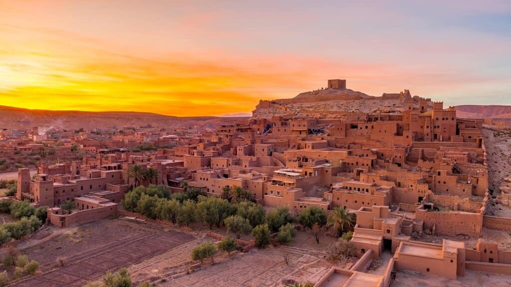 Panoramic view of the ancient earthen clay buildings of Ait Benhaddou kasbah built into a hillside in Morocco, illuminated by a vibrant orange and yellow sunset sky with a line of green trees and fields in the foreground.
