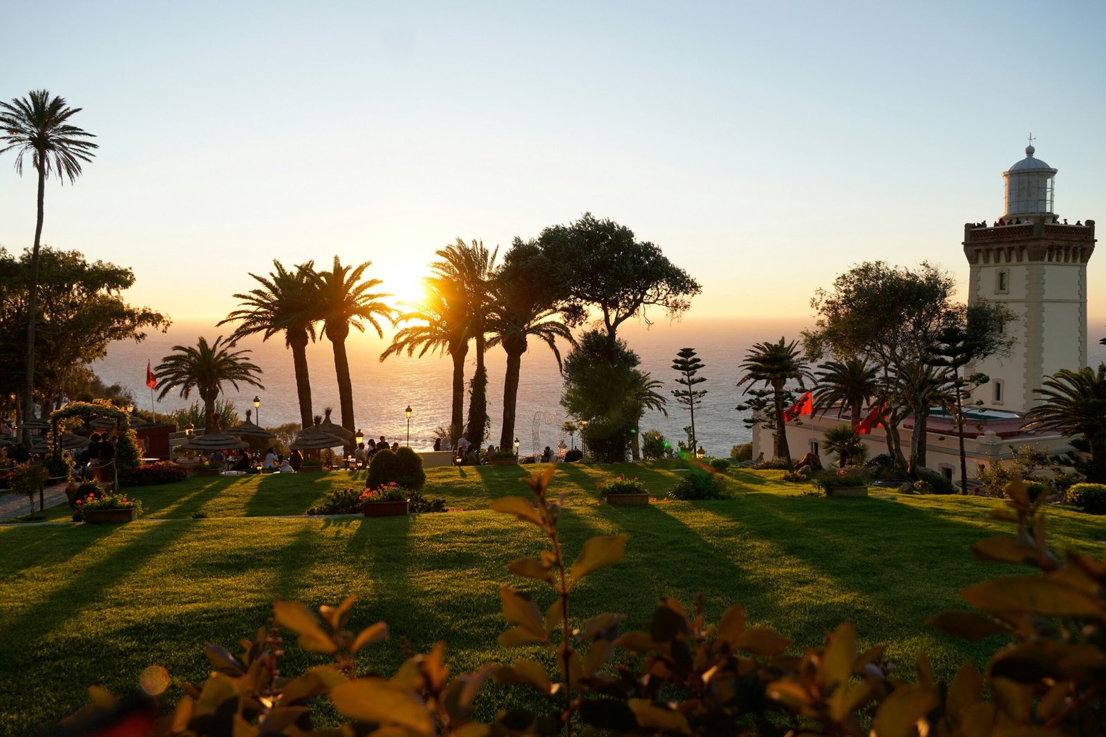 Cap Spartel Lighthouse Sunset in Tangier | Private Tours of Morocco - Rafia Morocco Tours - Rafia Morocco Tours A high-angle view of the historic Cap Spartel lighthouse at sunset near Tangier, with towering palm trees, a lush green lawn, and tourists enjoying tea overlooking the Atlantic Ocean on a private tour of Morocco.