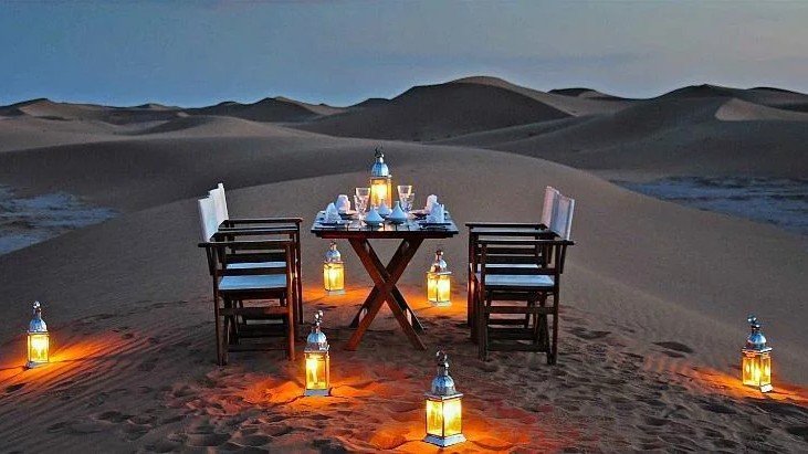 Luxury Desert Dining Experience | Private Tours of Morocco - Rafia Morocco Tours - Rafia Morocco Tours A private luxury dinner setup on a Sahara desert sand dune at dusk, featuring a set wooden table and white folding chairs surrounded by glowing silver lanterns, arranged for an exclusive private tour of Morocco.