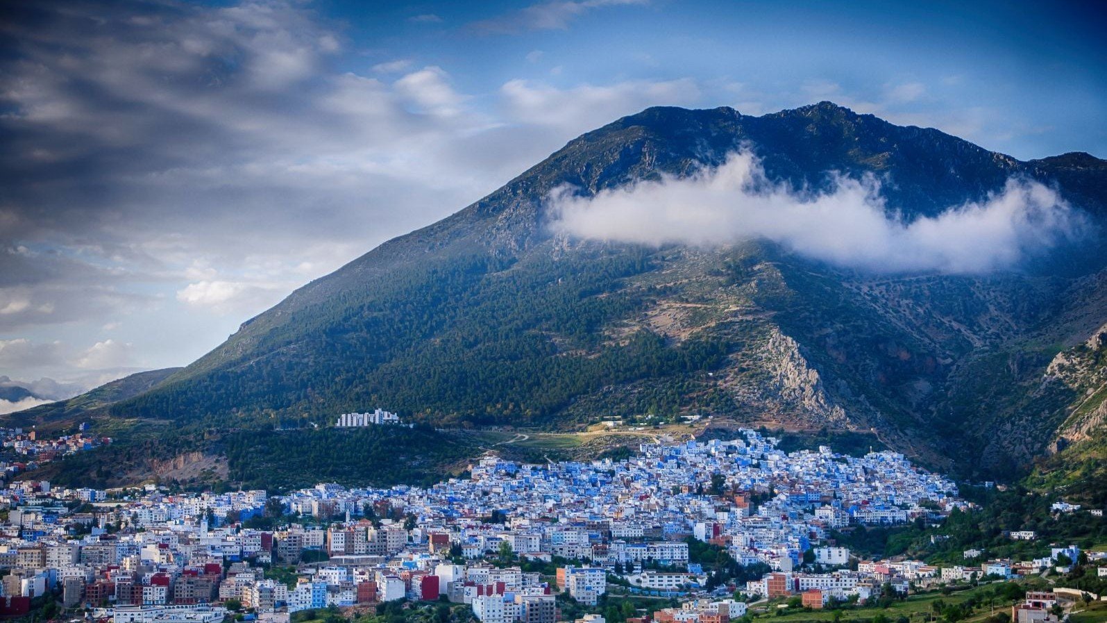 Panoramic View of Chefchaouen, the Blue City of Morocco - Rafia Morocco Tours - Rafia Morocco Tours A sweeping panoramic view of the blue-washed city of Chefchaouen nestled at the base of a massive, green-covered mountain peak, with a dramatic white cloud clinging to the hillside under a partly cloudy sky.