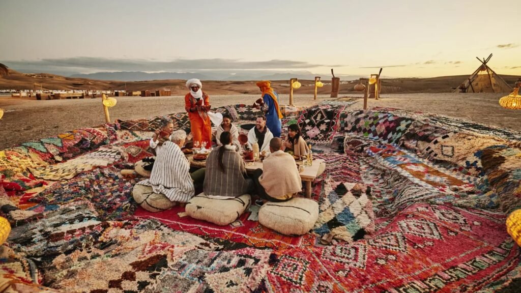 A group of travelers sitting on woven floor cushions around a low wooden dining table, entirely surrounded by layered colorful traditional Moroccan rugs in a desert camp setting, with hosts in traditional attire serving them under a pale sunset sky.