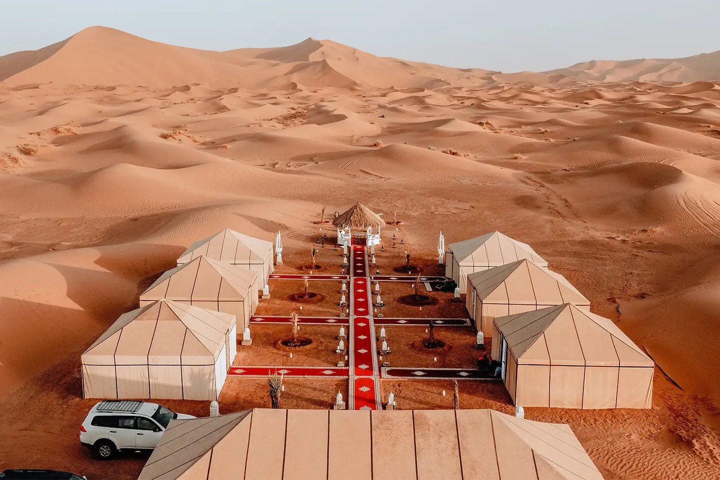 An aerial high-angle view of a luxury Sahara desert camp nestled among rolling orange sand dunes, featuring symmetric beige glamping tents arranged along a central red carpet pathway, captured during a private tour of Morocco.