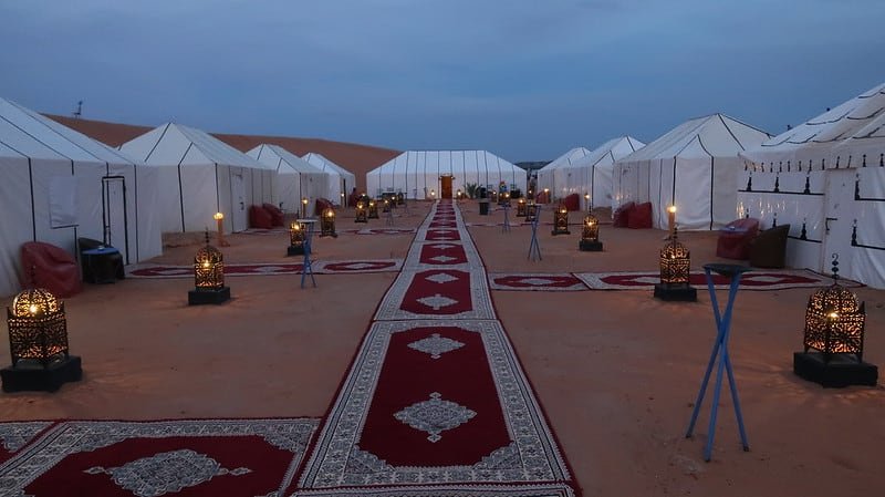 Luxury Desert Glamping Camp at Twilight, Sahara Morocco - Rafia Morocco Tours - Rafia Morocco Tours A long red Moroccan carpet walkway stretching through a luxury desert camp at twilight, lined with traditional black metal lanterns glowing with warm light, leading between white glamping tents pitched on the sand under a dark evening sky.