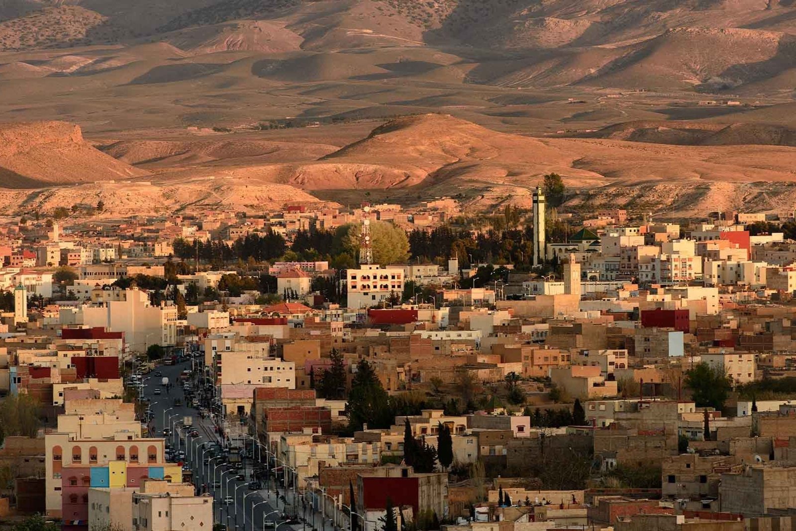 Gateway to the Sahara Desert | Private Tours of Morocco - Rafia Morocco Tours - Rafia Morocco Tours High-angle panoramic view of a Moroccan desert gateway town with flat-roofed buildings and a mosque minaret nestled at the base of the rugged, sun-drenched Atlas Mountains, captured during a private tour of Morocco.