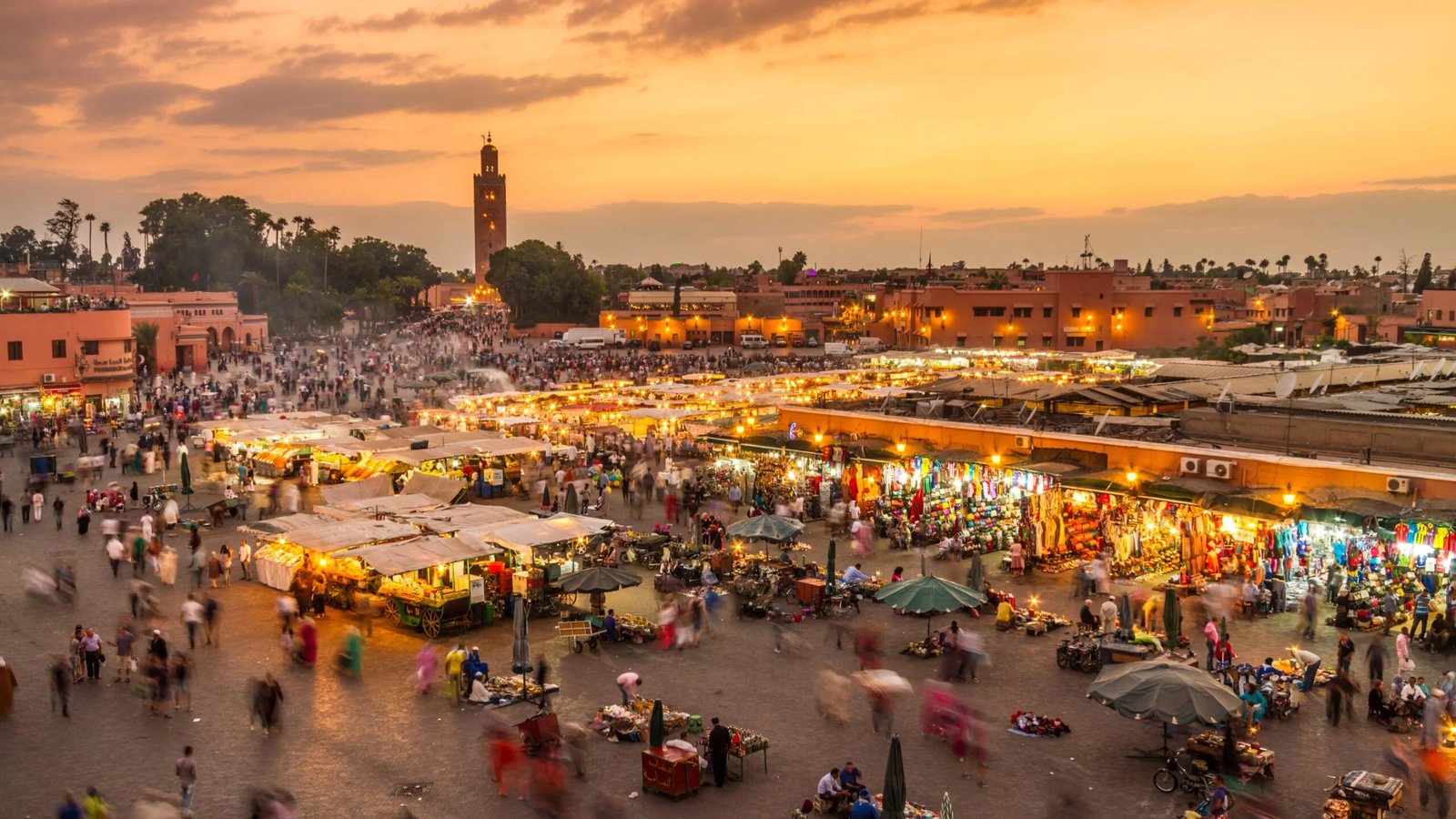 Jemaa el-Fnaa Square at Sunset, Marrakech - Rafia Morocco Tours - Rafia Morocco Tours High-angle panoramic view of the bustling Jemaa el-Fnaa square in Marrakech at twilight, filled with crowded crowds of people moving between illuminated food stalls and market canopies, with the Koutoubia Mosque minaret visible under a warm orange sunset sky.