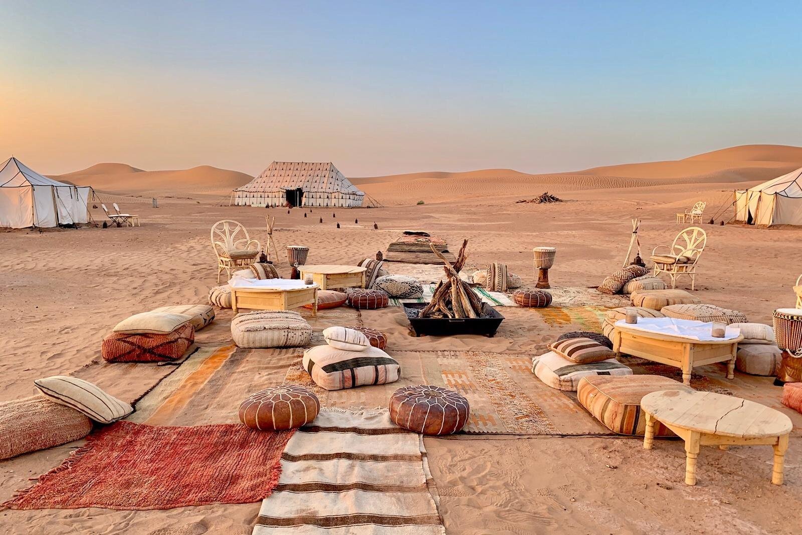 An outdoor lounge setup with traditional Moroccan rugs and cushions around a fire pit at a luxury Sahara desert camp during a private tour of Morocco.
