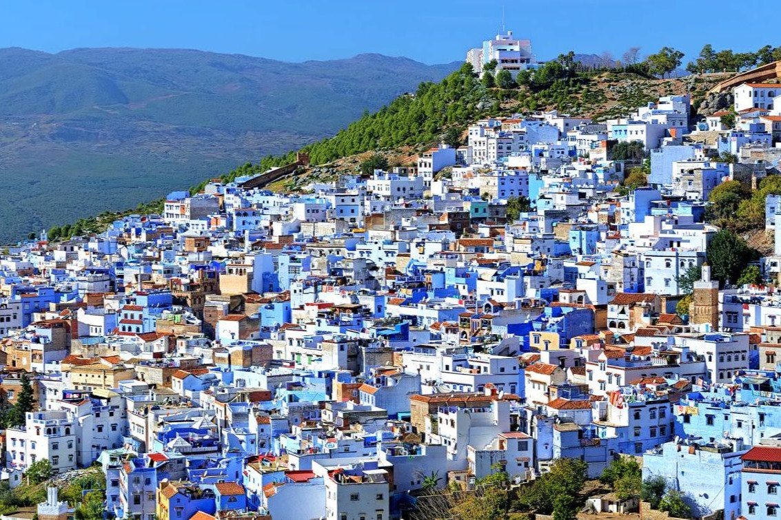 High-angle wide shot of the iconic blue-washed houses and traditional buildings of the Chefchaouen medina climbing the green slopes of the Rif Mountains under a clear blue sky, explored on a private tour of Morocco.
