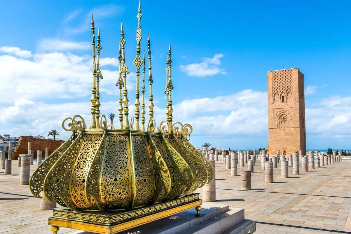 Hassan Tower Monument in Rabat | Private Tours of Morocco - Rafia Morocco Tours - Rafia Morocco Tours An ornate golden brass filigree lantern sculpture in the foreground at the historic Hassan Tower complex in Rabat, showcasing ancient stone pillars and the iconic unfinished Almohad minaret under a bright blue sky, visited on a private tour of Morocco.