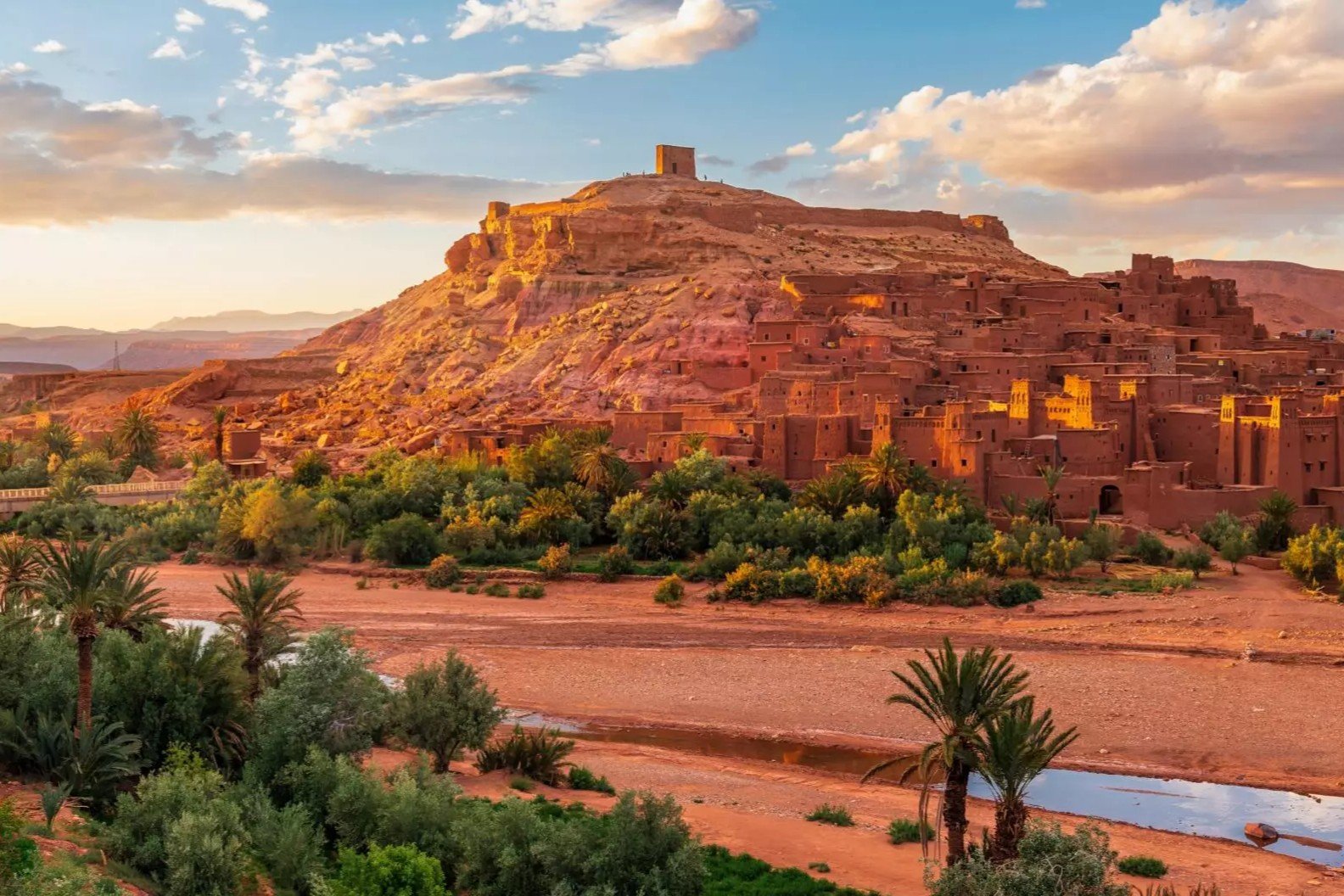 Ait Benhaddou Kasbah at Sunset | Private Tours of Morocco - Rafia Morocco Tours - Rafia Morocco Tours Panoramic wide shot of the fortified earthen clay ksar of Ait Benhaddou glowing in the golden sunset light, with green palm trees and a dry riverbed in the foreground, visited on a private tour of Morocco.