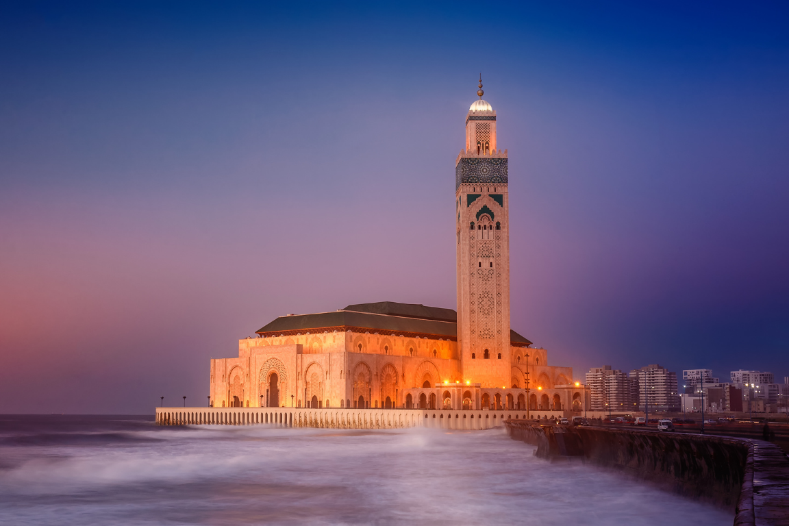 The illuminated Hassan II Mosque standing majestically on the Atlantic Ocean coast in Casablanca at twilight, featuring its towering minaret and detailed Moorish architecture under a purple sky, visited on a private tour of Morocco.