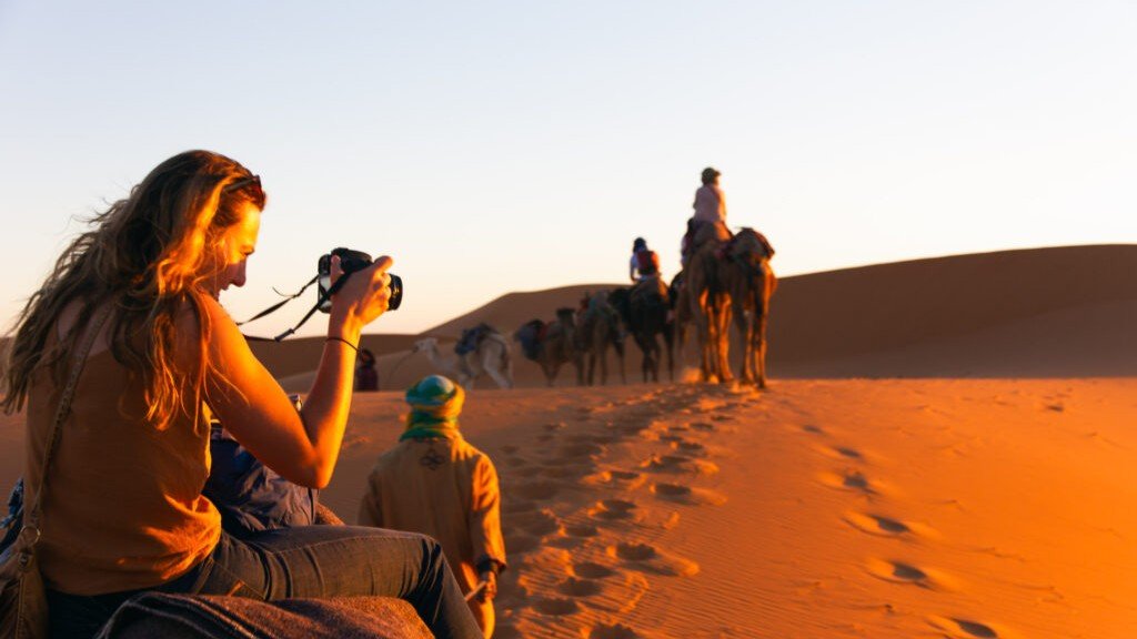 Capturing Memories on a Sahara Desert Tour | Private Tours of Morocco - Rafia Morocco Tours - Rafia Morocco Tours A female traveler on a private tour of Morocco sits on a camel holding a camera to photograph a camel caravan trekking ahead of her across rolling orange sand dunes in the Sahara Desert at sunset.