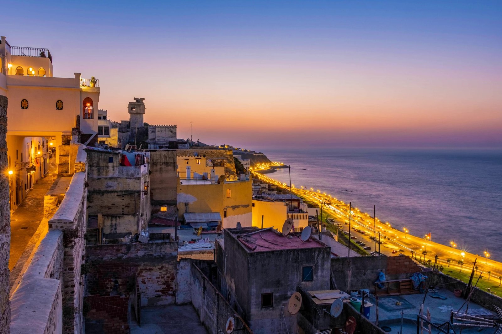 High-angle panoramic view of the historic Tangier medina rooftops at sunset, overlooking a brightly lit coastal highway stretching along the Atlantic ocean coast, captured during a customized private tour of Morocco.