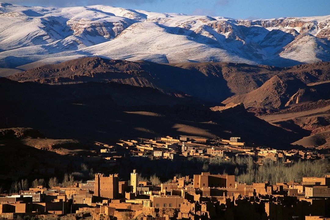 A high-angle panoramic view of traditional earthen clay kasbahs and villages in a mountain valley, contrasted against the massive, snow-covered peaks of the High Atlas Mountains, explored during a private tour of Morocco.