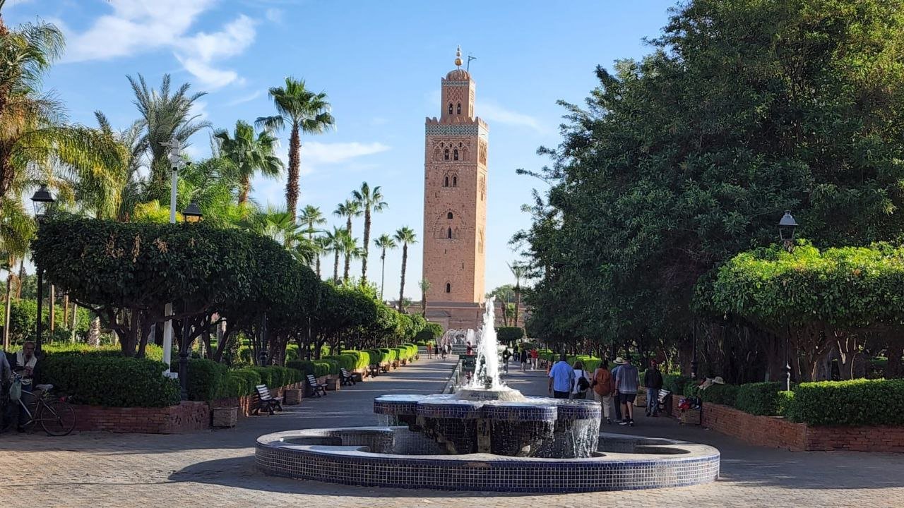 A scenic view of the tall sandstone Koutoubia Mosque minaret towering over Lalla Hasna Park in Marrakech, featuring a tiled water fountain in the foreground and a tree-lined walkway with palm trees under a bright blue sky.